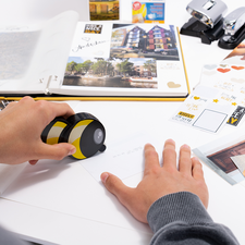 Eine Hand hält ein gelbes und schwarzes Gerät über ein Blatt Papier, umgeben von verschiedenen Dokumenten, Fotos und Bürobedarf.