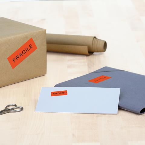 A box with the label 'FRAGILE,' next to an envelope marked 'URGENT!' and a gray folder on a table. Scissors and rolls of paper are also visible.