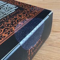 Cosmetic solutions: Black packaging with decorative orange patterns and text. The flap of the packaging is sealed with a transparent closure label.