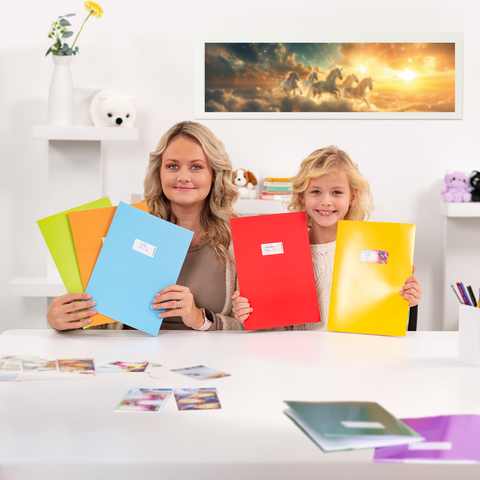 Two girls are sitting at a table holding colorful binders. On the table, there are photos, and in the background, there is a picture of unicorns.