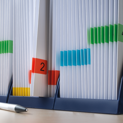 Kartonbox auf einem Tisch, mit einem roten Heftchen oben und einem grünen "TOP"-Schild. Im Hintergrund sind Bücherregale sichtbar.