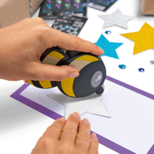 A hand uses a stamp to print a star on a piece of paper, surrounded by colorful stars and decorations.