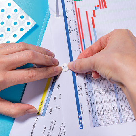 Hands attach a white snap button to a document in a binder, while a chart and tables are visible in the background.