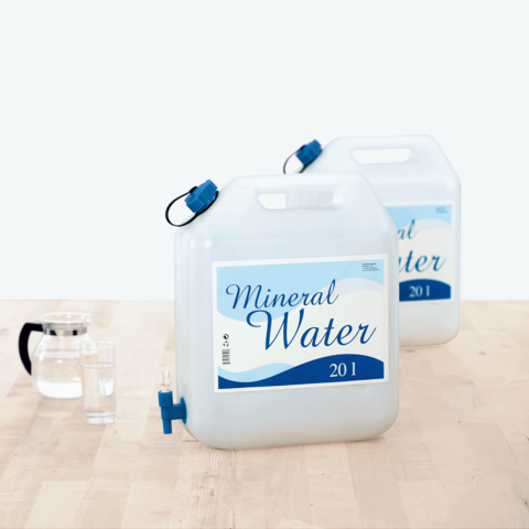Zwei 20-Liter-Behälter mit Mineralwasser, einer mit einem Auslaufhahn, neben einem Krug und einem Glas auf einem Tisch.