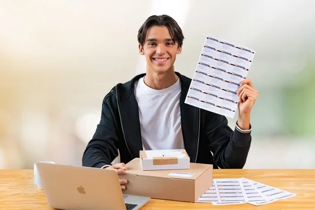 A young man is sitting at a table with a laptop, holding a sheet of stickers in his hand.