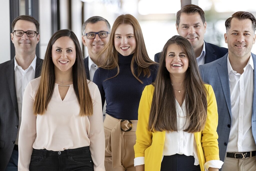 A group of seven people stands in a hallway, smiling and dressed in formal attire, some wearing blouses and suits.