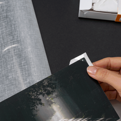 Eine Person hält ein Foto und fügt es in ein Fotoalbum ein, während sich daneben eine Verpackung mit Fotoecken befindet.