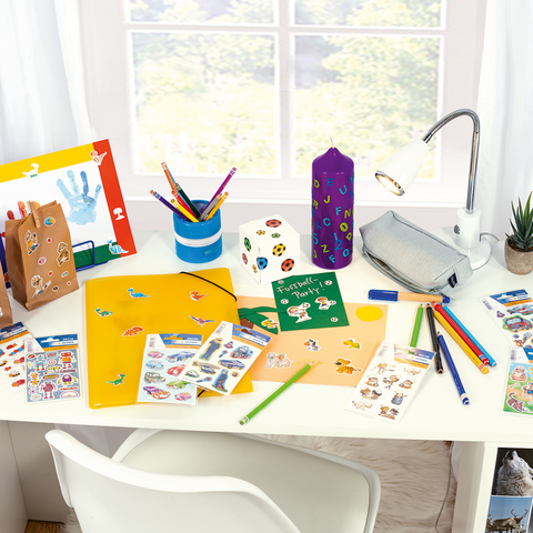 Bastelmaterialien auf einem Tisch, einschließlich Papier, Aufkleber, Stifte und eine Pflanze, mit Fenstern im Hintergrund.