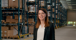 A woman stands in a warehouse with tall shelves full of boxes. She is wearing a black blazer and smiling.