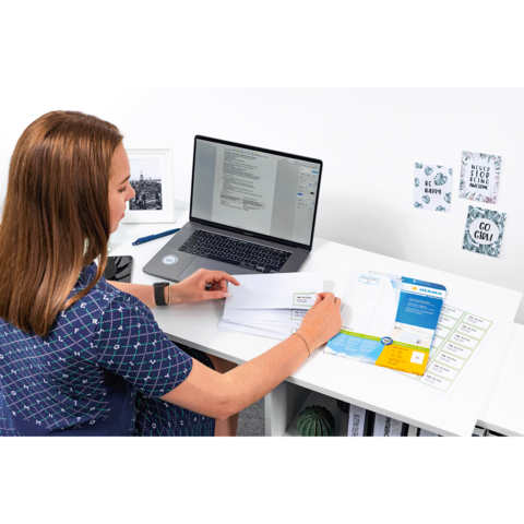 Eine Frau sitzt an einem Schreibtisch und arbeitet am Laptop, während sie Dokumente und Briefe sortiert.