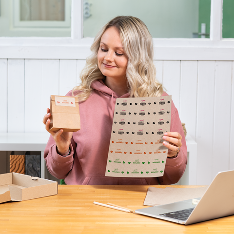 Eine Frau mit blonden, lockigen Haaren hält eine kleine Box und ein Blatt mit Aufklebern in der Hand, während sie an einem Tisch sitzt.