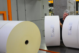 A worker in a printing press checks large rolls of paper while standing at a machine.