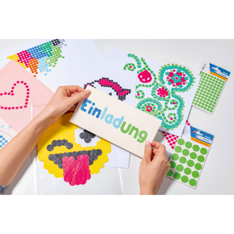 Hände halten eine Einladungskarte mit dem Text "Einladung", umgeben von bunten Bastelmaterialien und Aufklebern.