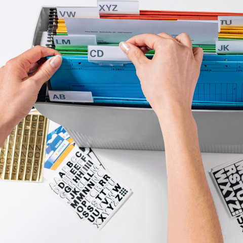 A hand is sorting colored tabs in a binder, while labels with letters are lying next to it.
