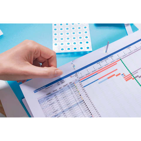 Eine Hand hält ein Blatt Papier mit einem Zeitplan oder einer Tabelle, während sich im Hintergrund ein Blatt mit Punkten befindet.