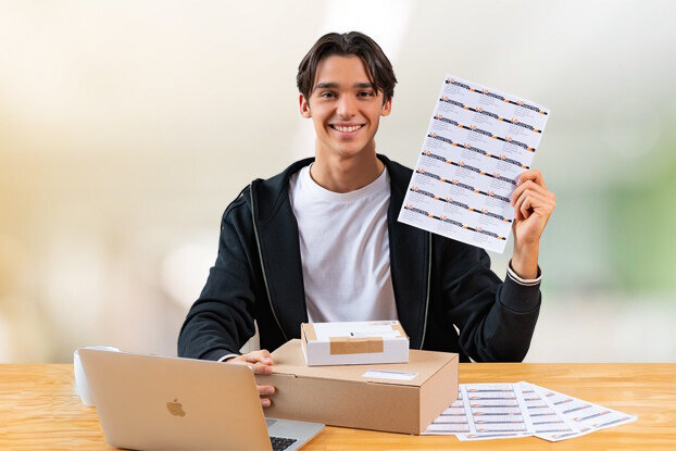 A young man is sitting at a table with a laptop, holding a sheet of stickers in his hand.
