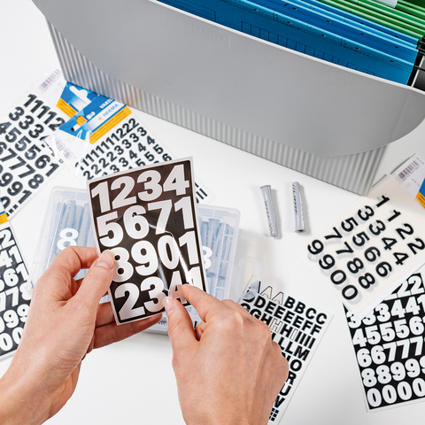 Hands hold a board with large numbers, surrounded by various letter and number stickers in a storage box.