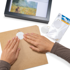 Hände drücken einen weißen Klebepunkt auf eine braune Pappe, während ein gerahmtes Bild und eine Packung mit weiteren Klebepunkten sichtbar sind.