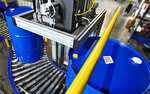 An automated conveyor system transports blue barrels in a storage environment, while a robotic arm is positioned above the barrels.