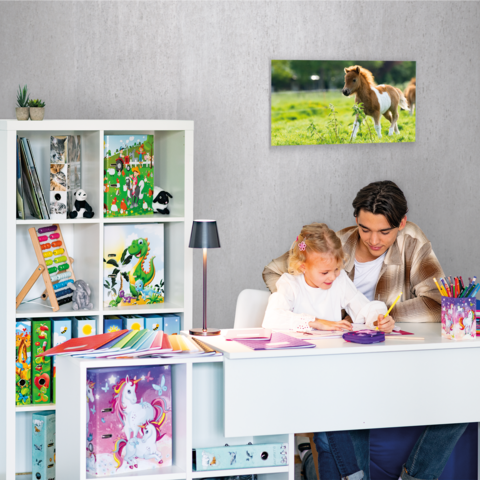 Ein Kind und ein Jugendlicher sitzen an einem Schreibtisch und zeichnen. Im Hintergrund steht ein Bücherregal und ein Bild eines Pferdes.