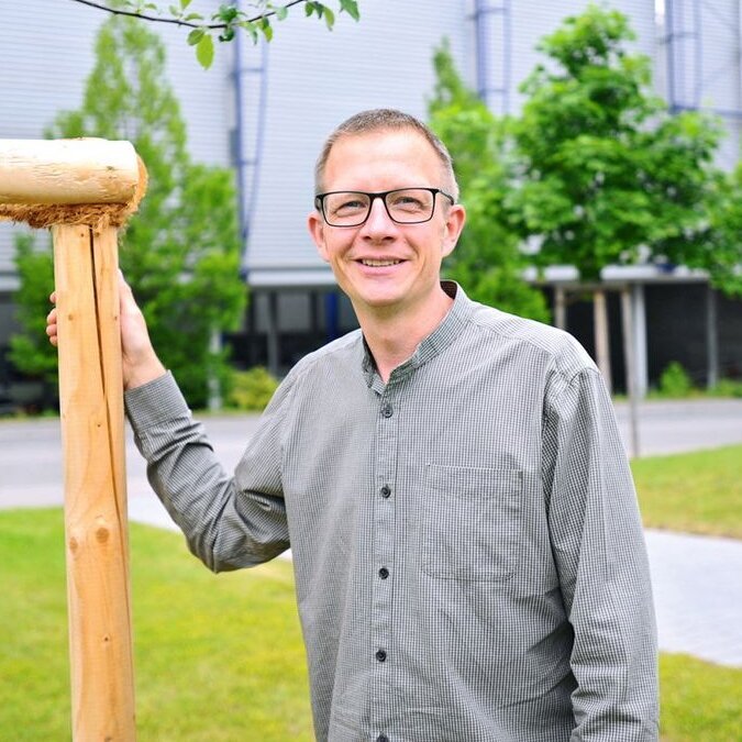 A man with glasses is smiling next to a wooden post in a green outdoor area with trees and modern buildings in the background.
