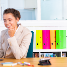 Eine Frau in einem Büro sitzt am Tisch und schaut auf einen Laptop. Im Hintergrund sind bunte Ordner sichtbar.
