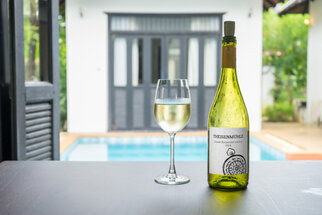 A bottle of white wine with a glass on a table, a pool in the background, and a bright, modern room.