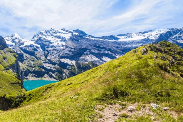 Panoramic view of green hills and snow-capped mountains, surrounded by clear water and a blue sky.