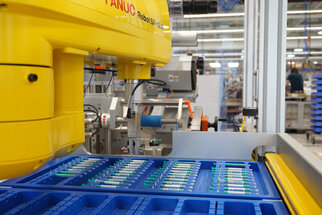 FANUC industrial robots picking parts from blue containers. In the background, additional machines and workstations are visible.