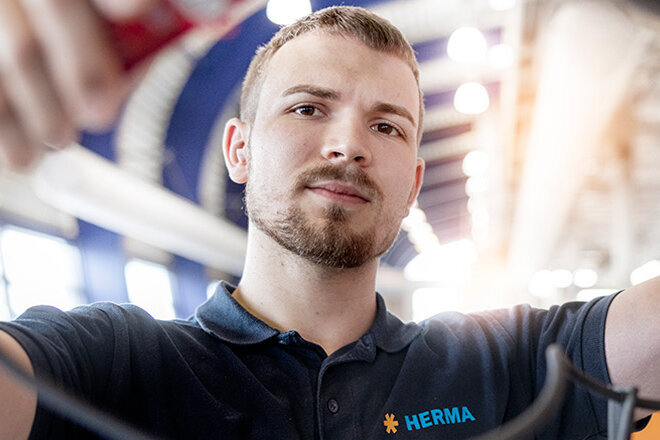 A young man in work clothes is holding cables in a modern setting with a building and trees in the background.