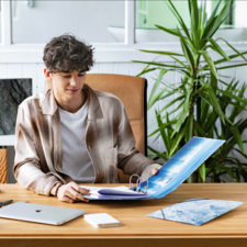 Ein junger Mann sitzt an einem Schreibtisch, schaut auf einen Ordner und arbeitet an einem Laptop. Pflanzen sind im Hintergrund sichtbar.