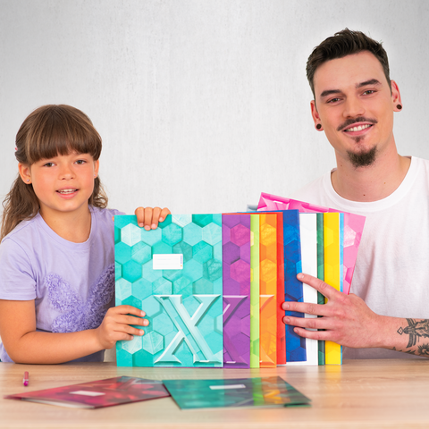 Ein Kind und ein Mann halten bunte, geometrisch gestaltete Ordner und zeigen sie in die Kamera. Auf dem Tisch liegen weitere Ordner.