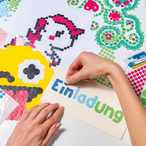 Eine Hand platziert bunte Punkte auf ein Einladungskarten-Design, um ein kreatives Kunstwerk zu gestalten. Mehrere Bastelmaterialien sind sichtbar.