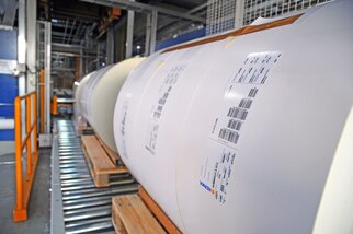 Rolls of paper on a conveyor system, each labeled with information and barcodes.