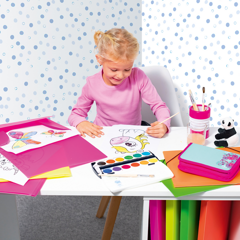 A child with blonde curls sits at a table, painting with watercolors. Colorful drawings and craft materials are scattered around her.