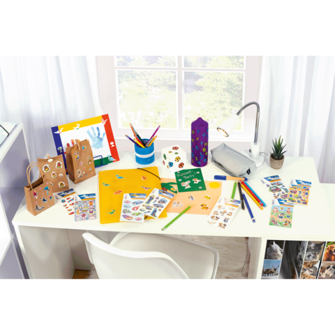 Bastelmaterialien auf einem Tisch, einschließlich bunter Stifte, Aufkleber, Papier und einer Pflanze, mit Fenstern im Hintergrund.