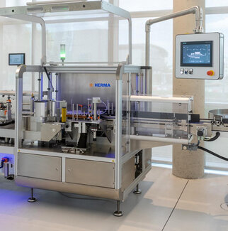Automated labeling machine from HERMA with a control panel and various mechanical components for labeling products.