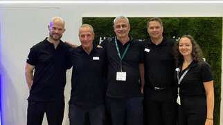 Five people in black clothing are standing side by side in front of a wall featuring the HERMA logo and the slogan 'Coating your success.