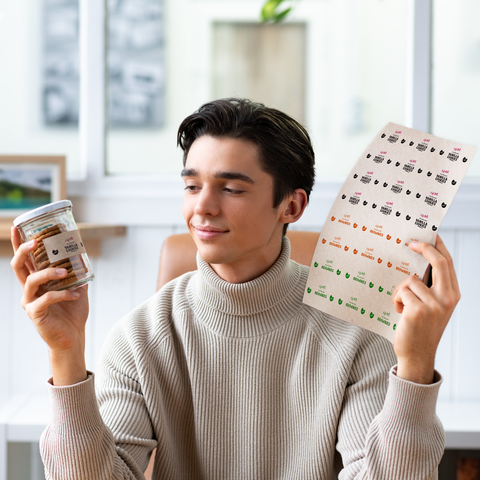 A young man is holding a glass of snacks in one hand and a label with various designs in the other hand.