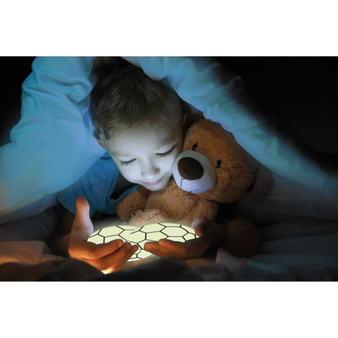 Ein Kind sitzt im Bett, umgeben von einer Decke, und hält ein leuchtendes Objekt in den Händen, während es einen Teddybären umarmt.