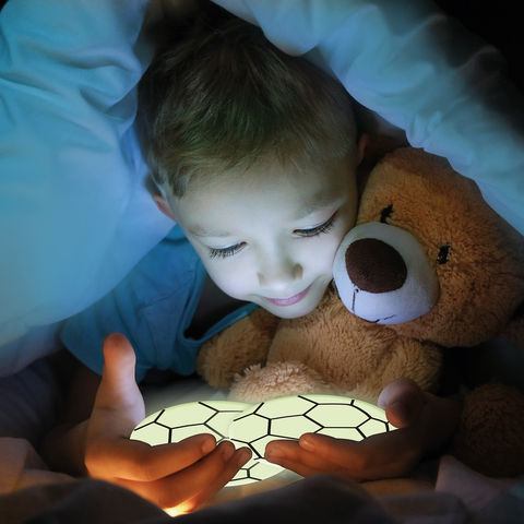 Ein Kind sitzt im Bett, umgeben von einer Decke, und hält ein leuchtendes Objekt in den Händen, während es einen Teddybären umarmt.