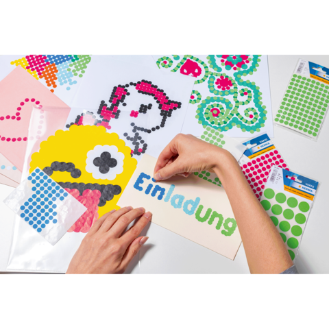 Bunte Bastelmaterialien liegen auf einem Tisch, während eine Hand einen Aufkleber auf eine Einladung mit dem Wort "Einladung" platziert.