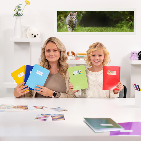 A woman and a girl are holding colorful binders while sitting at a table with photos. In the background, a cat can be seen.