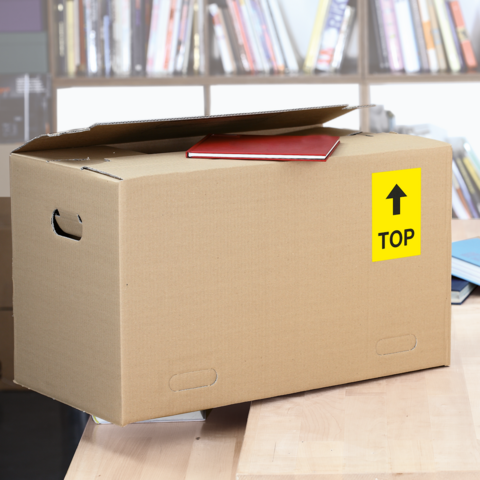 A cardboard box with a red notebook on top, labeled with a yellow "TOP" sign, sits on a table in a library.