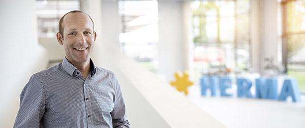 A man in a checked shirt smiles in a modern office with the sign "HERMA" in the background.